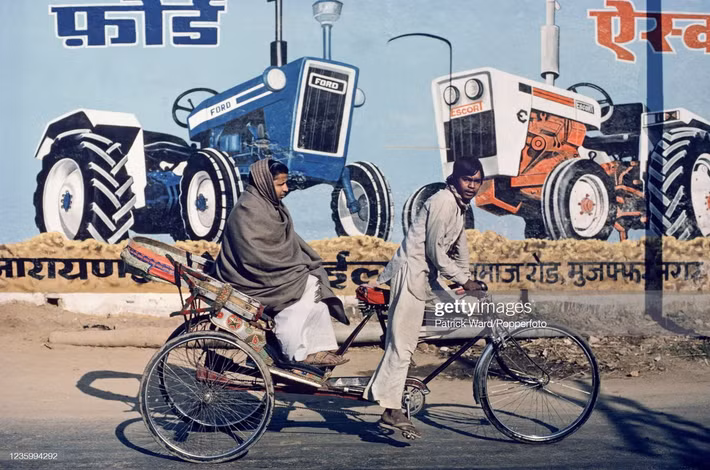 Đôi nam nữ di chuyển bằng xe đạp ba bánh ngang qua pa-nô quảng cáo xe công nông của hãng Ford ở thành phố Delhi, Ấn Độ năm 1970. Ảnh: Patrick Ward/ Getty Images.
