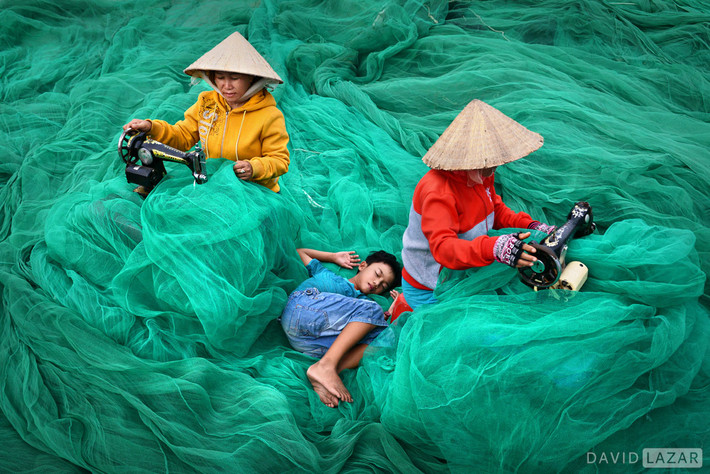 Cậu bé ngủ trên tấm lưới cá đang được vá bởi hai phụ nữ tại một làng chài Nam Trung Bộ.