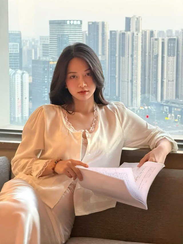 Trong khung hình, hot girl ngồi bên cửa kính, phía sau là hoàng hôn rực rỡ. Bộ váy lụa trắng kem cùng bó hoa tươi tạo nên vẻ đẹp sang trọng và tinh tế.