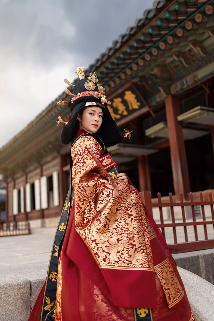 Đứng trước cánh cửa cung điện, nữ diễn viên khoác lên mình bộ lễ phục đỏ thêu hoa văn vàng nổi bật. Dáng đứng ngay ngắn, hai tay đặt trước người cùng vẻ mặt nghiêm nghị của cô khiến khung hình gợi nhớ đến những cảnh trong phim cổ trang.