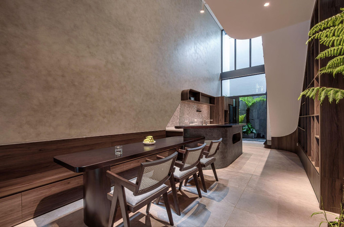 Bếp và bàn ăn được bố trí dọc nhà, sử dụng chất liệu gỗ tông trầm, kết hợp ánh sáng từ thông tầng phía sau tạo cảm giác ấm áp. Ảnh: LonTon Studio
