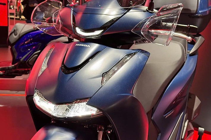 Honda SH125i/150i 2026 được tái thiết kế sâu sắc, lấy cảm hứng từ “đàn anh” SH350i. Phần đầu xe nổi bật với cụm đèn pha LED kép hoàn toàn mới, đi cùng dải đèn định vị sắc nét hơn. Cản trước được tinh chỉnh trở nên mềm mại và khí động học hơn, trong khi từng chi tiết nhỏ như sơn xe, ống xả hay vành đen bóng đều được hoàn thiện tỉ mỉ.